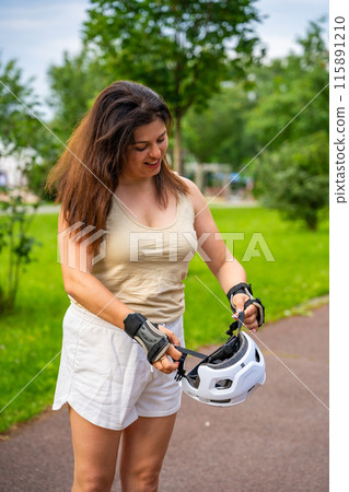 Portrait of roller skater woman putting on helmet in inline skates before rollerblading in park. Outdoor activities. Summer roller skating. 115891210