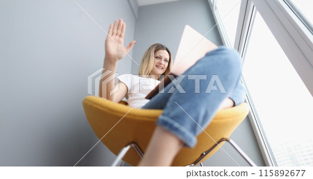 Young woman sitting on armchair and waving hand at laptop screen Young woman sitting on armchair and waving hand at laptop screen 115892677