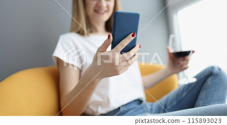 Woman holds smartphone and glass of red wine in her hands 115893023