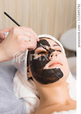Cosmetologist applying black sheet mask on woman's face in beauty salon. 115895792