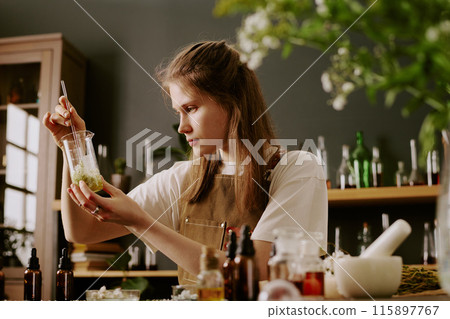 Young female perfumer sitting at work desk with perfume craft items and admiring at golden liquid with wildflowers petals while she mixing it Young female perfumer sitting at work desk with perfume craft items and admiring at golden liquid with wildflowers petals while she mixing it 115897767