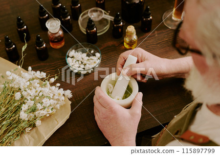 Mature perfumer crushing dried daisies in ceramic mortar with white ceramic pestle, he sitting at work desk with different essence in brown glass bottles Mature perfumer crushing dried daisies in ceramic mortar with white ceramic pestle, he sitting at work desk with different essence in brown glass bottles 115897799