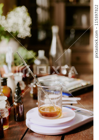 Crystal flask with caramel shade aromatic essence with glass pipette on plastic kitchen scale, old wooden table with perfume sample bottles and plants 115897822