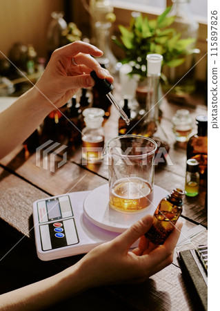 Medium close up of female hand holding brown glass bottle with aroma essence and glass pipette while she adding magnificent aromatic essence in crystal flask Medium close up of female hand holding brown glass bottle with aroma essence and glass pipette while she adding magnificent aromatic essence in crystal flask 115897826