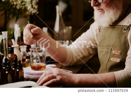 Senior perfumer sitting at table with brown glass bottles of essential oils and mixing cognac shade essences with help of long glass stick 115897839