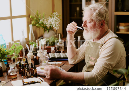 Mature perfumer smelling new handmade scent with help of blotter while he sitting at table with dozens of perfume equipment 115897844