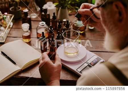 Over shoulder shot of mature perfumer adding extra essential oil while glass flask with perfume sample on kitchen scales according to his notes 115897856