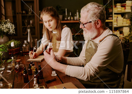 Mature perfumer showing his brand new sample of unique perfume while his granddaughter admiring it and making personal notes 115897864