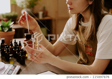 Medium close up of female perfumer adding new essential oil while she preparing new perfume for selling 115897876