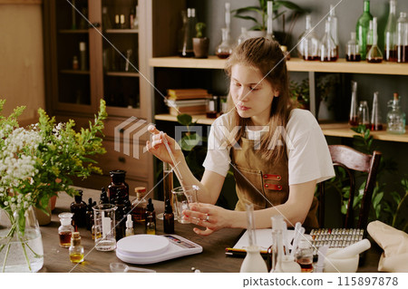 Female perfumer sitting at table and creating new scent while she mixing essential oils Female perfumer sitting at table and creating new scent while she mixing essential oils 115897878