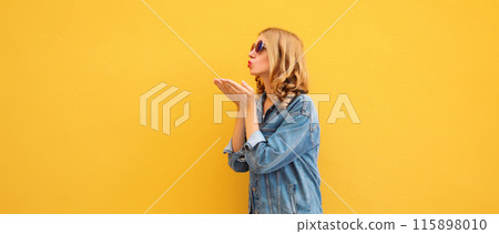 Beautiful stylish young woman blowing kiss in glasses, denim jacket posing on yellow wall background 115898010
