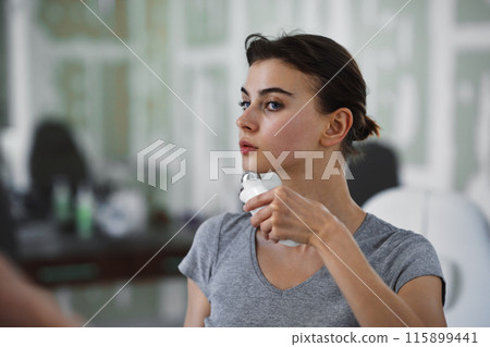 A young woman massages her face with a microcurrent gadget while looking in the mirror. A young woman massages her face with a microcurrent gadget while looking in the mirror. 115899441