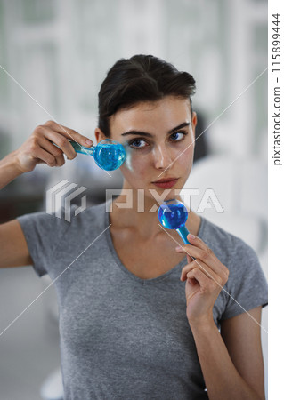 A young woman is massaging her face with blue glass balls while looking in the mirror. 115899444