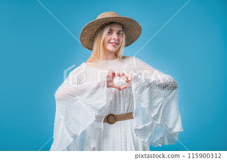 Smiling stylish teenager girl showing sign of shape heart. Positive blonde boho styled lady on blue 115900312