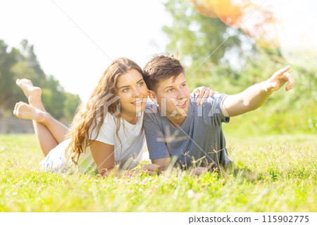 Happy couple lie on grass point with hand to something Happy couple lie on grass point with hand to something 115902775