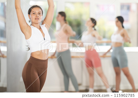 Portrait of cheerful active girl exercising ballet moves in fitness studio. Group of young ballet dancers in background near ballet barre Portrait of cheerful active girl exercising ballet moves in fitness studio. Group of young ballet dancers in background near ballet barre 115902785