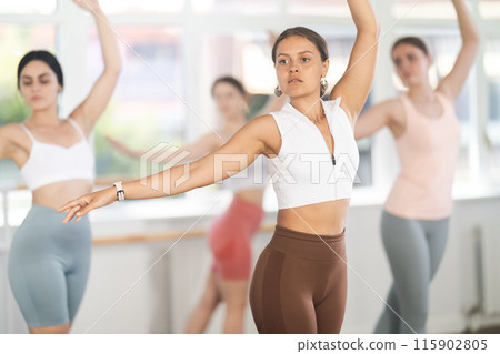 Diligent beautiful young female dancer exercising ballet moves during training with group of sporty people in training room Diligent beautiful young female dancer exercising ballet moves during training with group of sporty people in training room 115902805