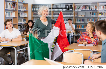 Mature female teacher shows the flag of Mexico to teenagers and talks about this country during lesson at library school Mature female teacher shows the flag of Mexico to teenagers and talks about this country during lesson at library school 115902907