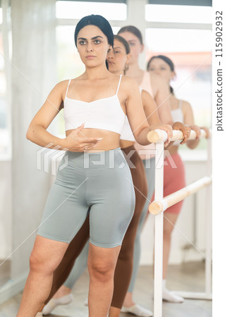 Positive young girl with pretty female engaged in group class perform exercise near ballet barre, standing in first position of ballet stance in dance studio 115902932