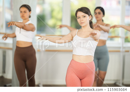 Cheerful slim active girl training various ballet poses during group classes with ballet dancers at barre in choreography studio 115903084