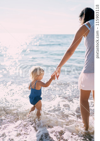 Little girl in a swimsuit stands in the sea waves holding her mom hand. High quality photo 115903118