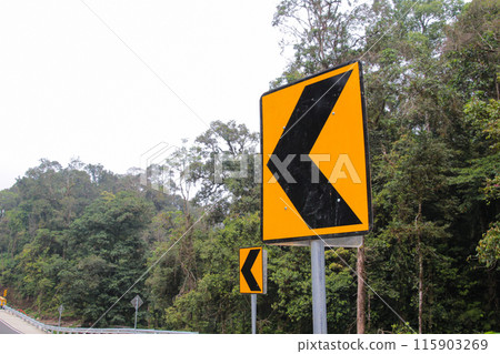 Yellow and Black Arrow Road Sign with Trees in the Background Yellow and Black Arrow Road Sign with Trees in the Background 115903269