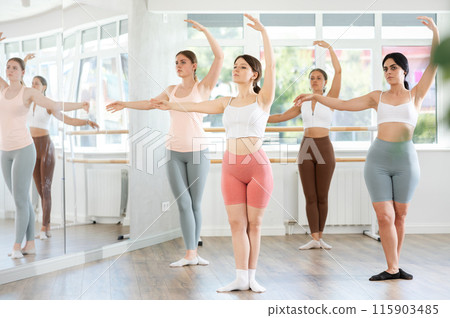 Sporty happy young girl demonstrating fourth ballet position with group of young girls in choreography studio Sporty happy young girl demonstrating fourth ballet position with group of young girls in choreography studio 115903485