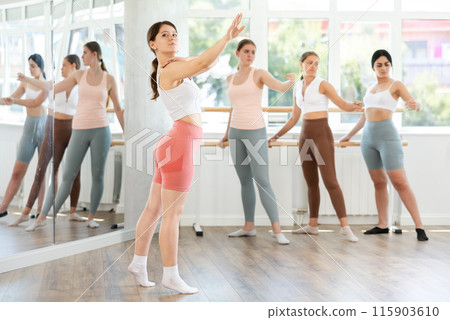 Concentrated positive young woman participating in ballet class, practicing basic movements in choreography studio. Group of young ballet dancers in background near ballet barre Concentrated positive young woman participating in ballet class, practicing basic movements in choreography studio. Group of young ballet dancers in background near ballet barre 115903610