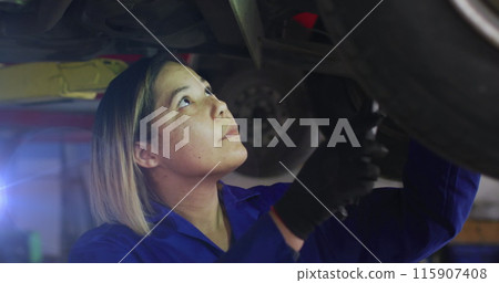 Image of asian female mechanic standing under car examining vehicle with hand tool in garage 115907408