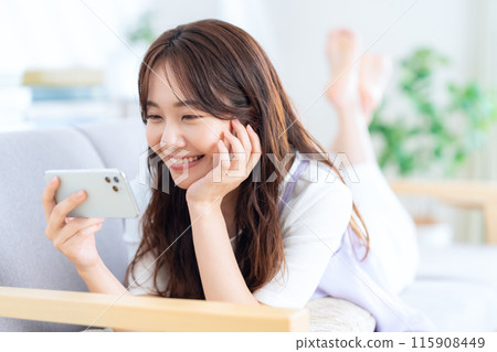 一位年輕女子在客廳一邊看著智慧型手機一邊放鬆 115908449