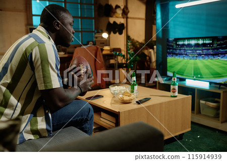 Young African American man holding ball sitting on couch in living room watching football game on TV in evening Young African American man holding ball sitting on couch in living room watching football game on TV in evening 115914939