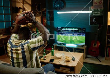 Unrecognizable young African American football fan holding ball above his head watching match on TV in living room, copy space Unrecognizable young African American football fan holding ball above his head watching match on TV in living room, copy space 115914944