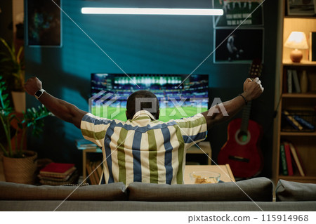 Back view of unrecognizable young Black man sitting on sofa in living room enjoying watching football match on TV 115914968