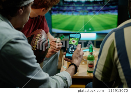 Crop shot of young male friends betting on team in app on smartphone while watching football match on TV Crop shot of young male friends betting on team in app on smartphone while watching football match on TV 115915070