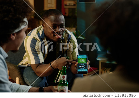 Young African American man showing hos friends how to bet on soccer team with use of app on smartphone 115915080