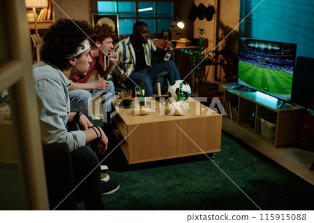 Group of multi-ethnic male friends sitting in living room watching soccer match on TV in evening, copy space Group of multi-ethnic male friends sitting in living room watching soccer match on TV in evening, copy space 115915088