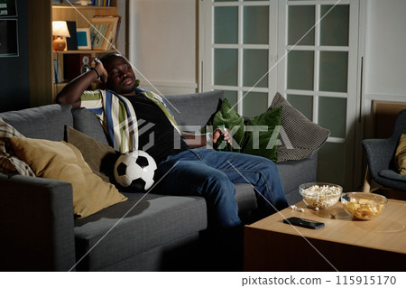 Young Black man holding bottle of beer sitting relaxed on sofa in living room watching soccer match on TV, copy space 115915170