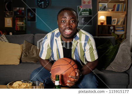 Medium shot of cheerful African American guy sitting on couch watching basketball match on TV late in evening Medium shot of cheerful African American guy sitting on couch watching basketball match on TV late in evening 115915177