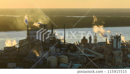 Huge factory with high chimneys polluting atmosphere with carbon dioxide smoke from production process at plant manufacturing yard. Industrial site at sunset 115915515