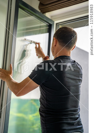 Young man cleans the windows in the house. He splashes detergent, rubs and removes water at the glass 115918983