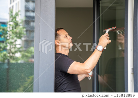 Young man cleans the windows in the house. He splashes detergent, rubs and removes water at the glass 115918999