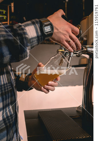 Bartender standing behind bar counter filling glass with white beer from tap 115919761