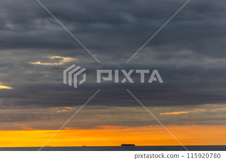 The sun is setting over the ocean with ships in the distance, under a dramatic sky 115920780
