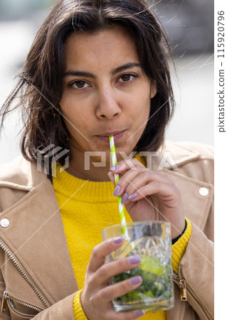 A woman in a yellow sweater enjoys a refreshing drink outdoors, sipping through a striped straw A woman in a yellow sweater enjoys a refreshing drink outdoors, sipping through a striped straw 115920796