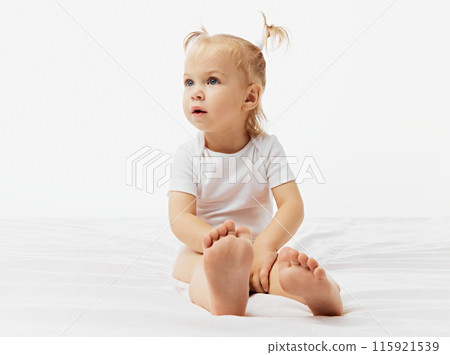 Curious kid, baby girl with blonde pigtails sits on white surface, dressed in onesie against white studio background. Curious kid, baby girl with blonde pigtails sits on white surface, dressed in onesie against white studio background. 115921539