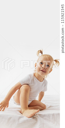 Poster. Blonde toddler with pigtails, wearing onesie sitting on bed against white studio background with negative space to insert your text. 115921545