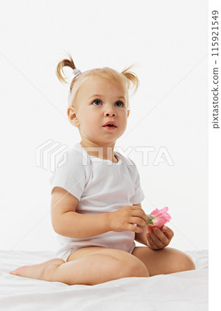 Curious child, baby girl sitting on white bed and looks upward holding pink flower against white studio background. 115921549