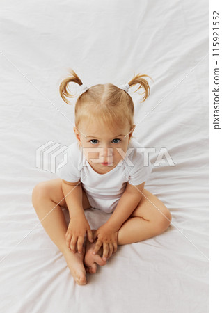 View from above serious-looking toddler with blonde hair styled into two small pigtails, sitting cross-legged against white studio background. 115921552