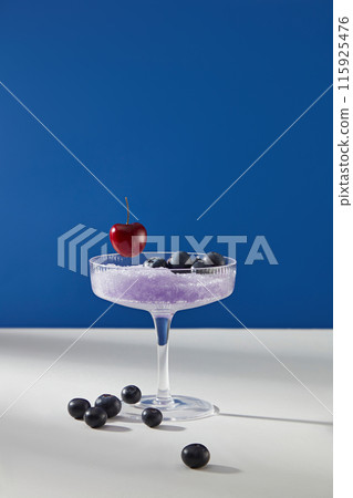 A cocktail glass containing purple grinding ice with some fresh blueberry and a red cherry decorated above, isolated on dark blue background. Blank space for designing and adding text 115925476