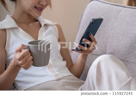Young Woman Relax on Couch Using Smartphone and Drinking Coffee for Social Media Browsing Young Woman Relax on Couch Using Smartphone and Drinking Coffee for Social Media Browsing 115927245